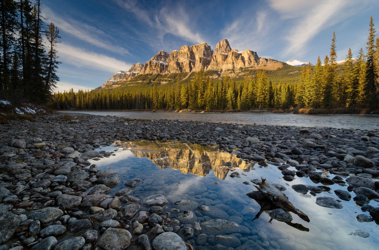 Castle Mountain catching low sun above a band of spruce, reflected in a still pool on the stony bank of the Bow River in Banff National Park.
