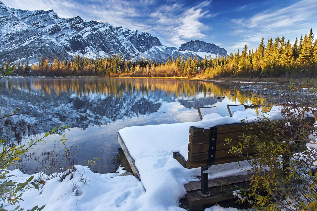 Snow-dusted Front Range peaks reflected in a pond at Many Springs in Bow Valley Provincial Park, with golden trees and a snowy bench in the foreground.