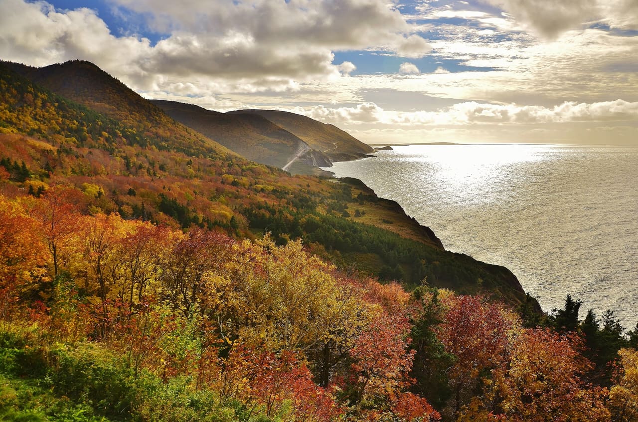 Headlands along the Cabot Trail in Cape Breton Highlands National Park covered in autumn colour above a sunlit sea.