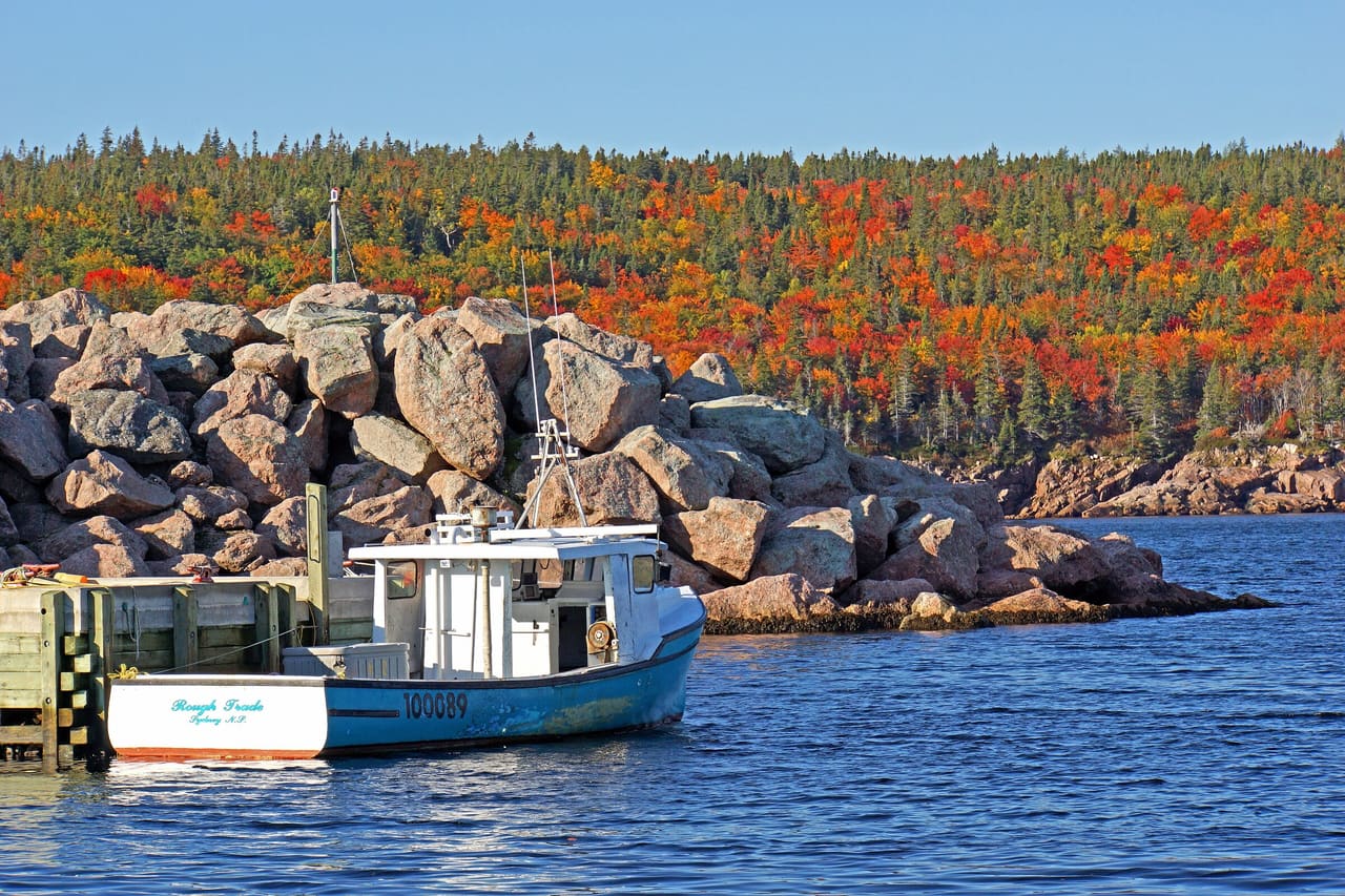 A fishing boat moored by a granite breakwater at Neil's Harbour, with hills of red and orange autumn trees behind.