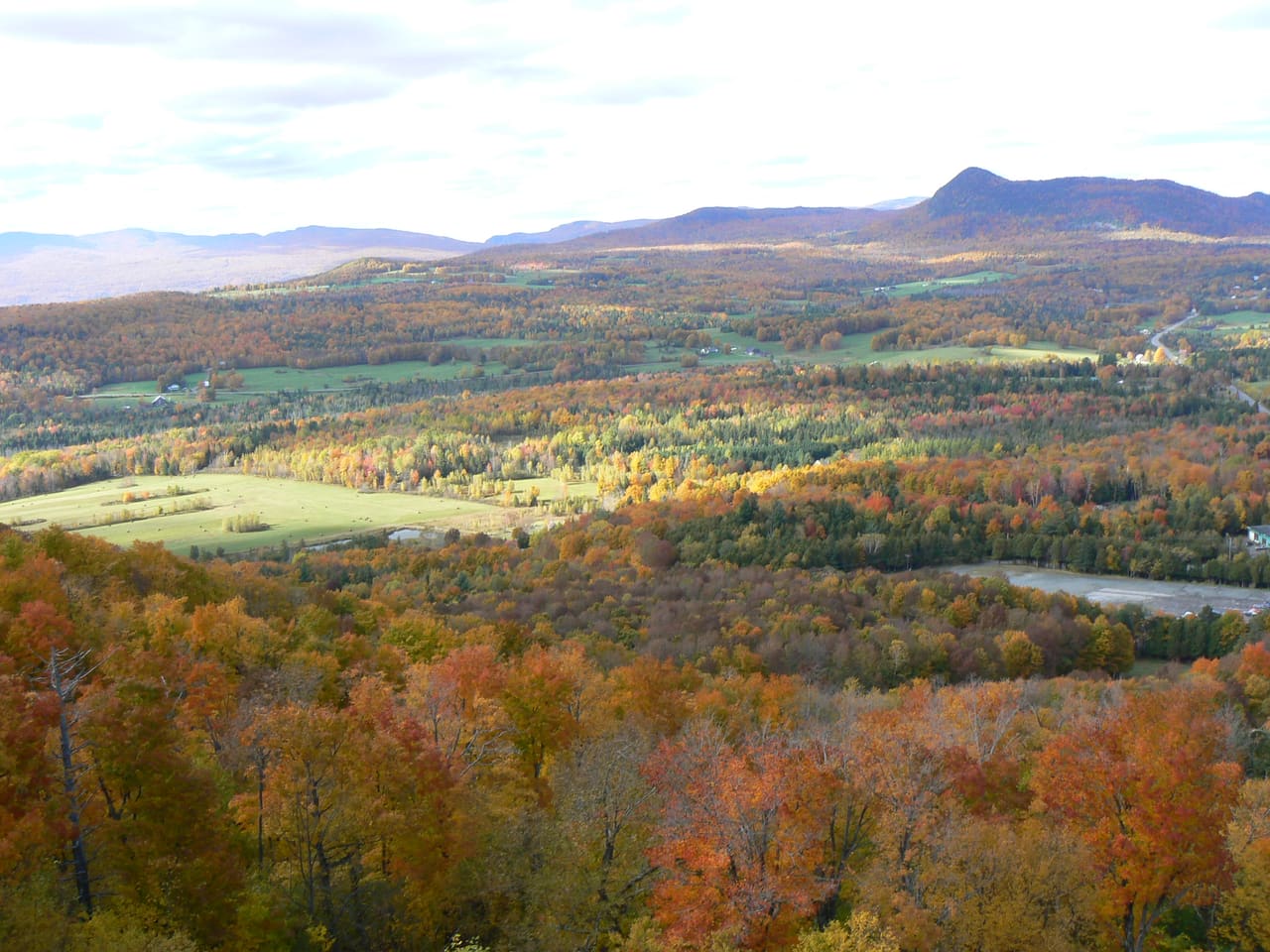 Autumn maple forest in red and orange below Owl's Head, with farm fields and rolling hills of the Eastern Townships stretching to the mountains on the horizon.