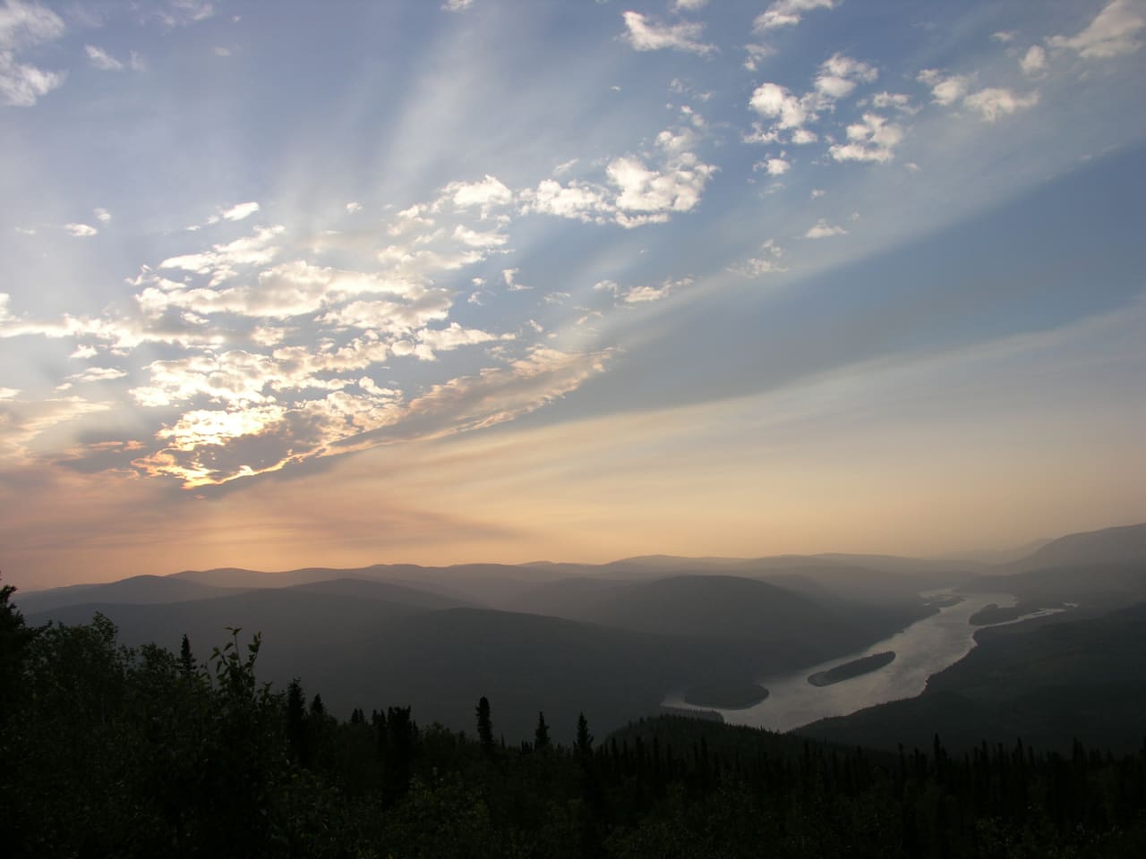 Evening sun rays over forested hills and the Yukon River winding through its valley, seen from the Midnight Dome above Dawson City.